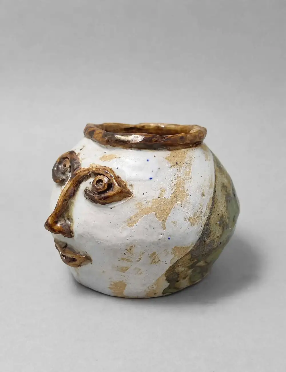 Round white ceramic pot with a face built into the clay, spiral eyes and amber glaze linework